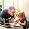 A dad and his young daughter writing in a notebook.