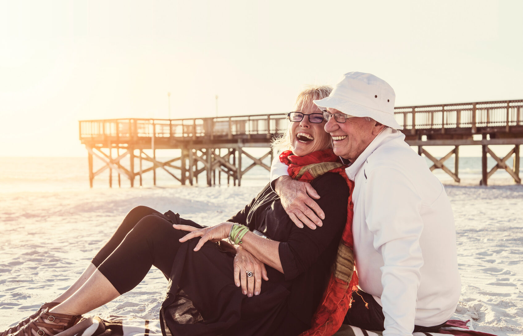 An older couple cuddles and smiles on a blanket by a pier