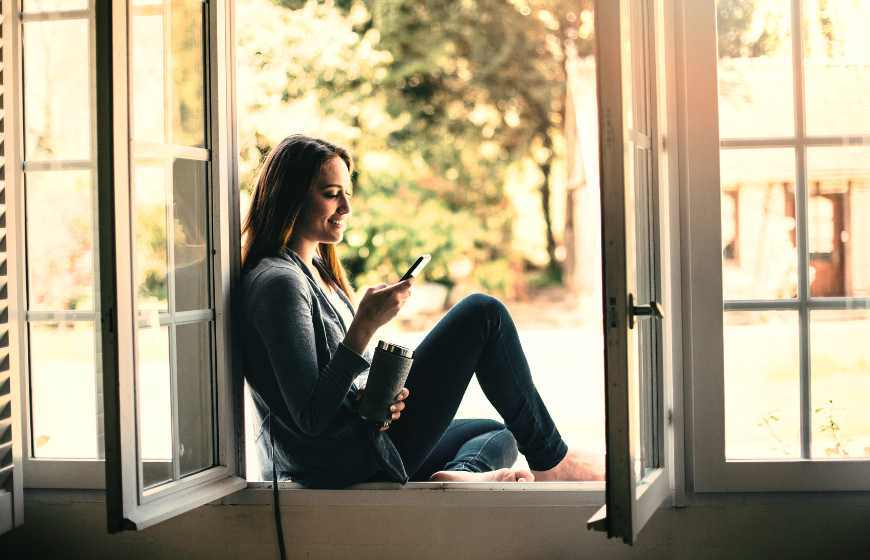 A woman sitting on the ledge of an open window, looking at her phone.
