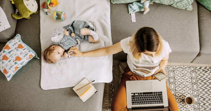 A mom sits on the floor with a laptop and a phone while holding one hand on her baby on the couch.