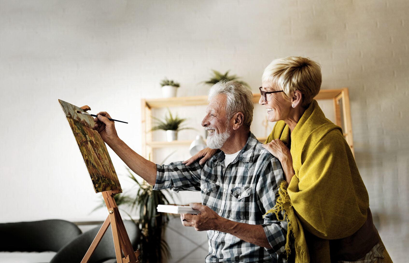 An older man smiling and painting with an older woman smiling and standing behind him.