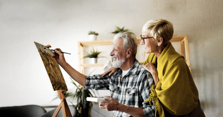 An older man smiling and painting with an older woman smiling and standing behind him.