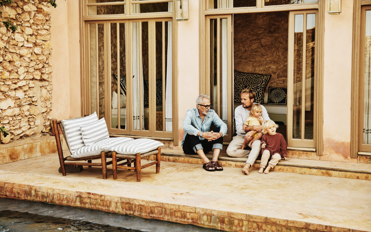 Father and adult son in discussion while relaxing with family on poolside terrace of villa